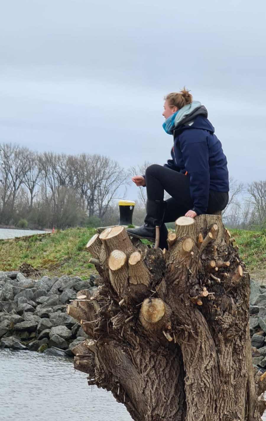 Marie sitzt nachdenklich auf einem Baumstumpf am Wasser